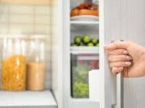 Woman's hand opening the door of a fridge to reveal produce with bins of dry goods on the counter in the background
