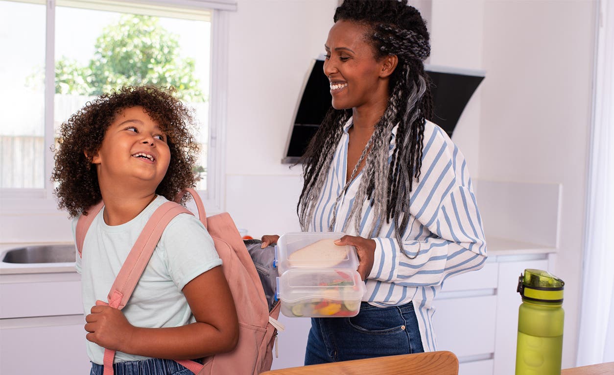 Lunchbox Tips with Dreena Burton A mother stands behind her daughter in the kitchen ready to put a bag of snacks into her daughters backpack. Both are smiling and happy.