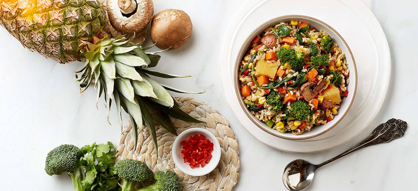 A bowl of No-Fry Fried Rice next to some raw mushrooms, broccoli, and a whole pineapple
