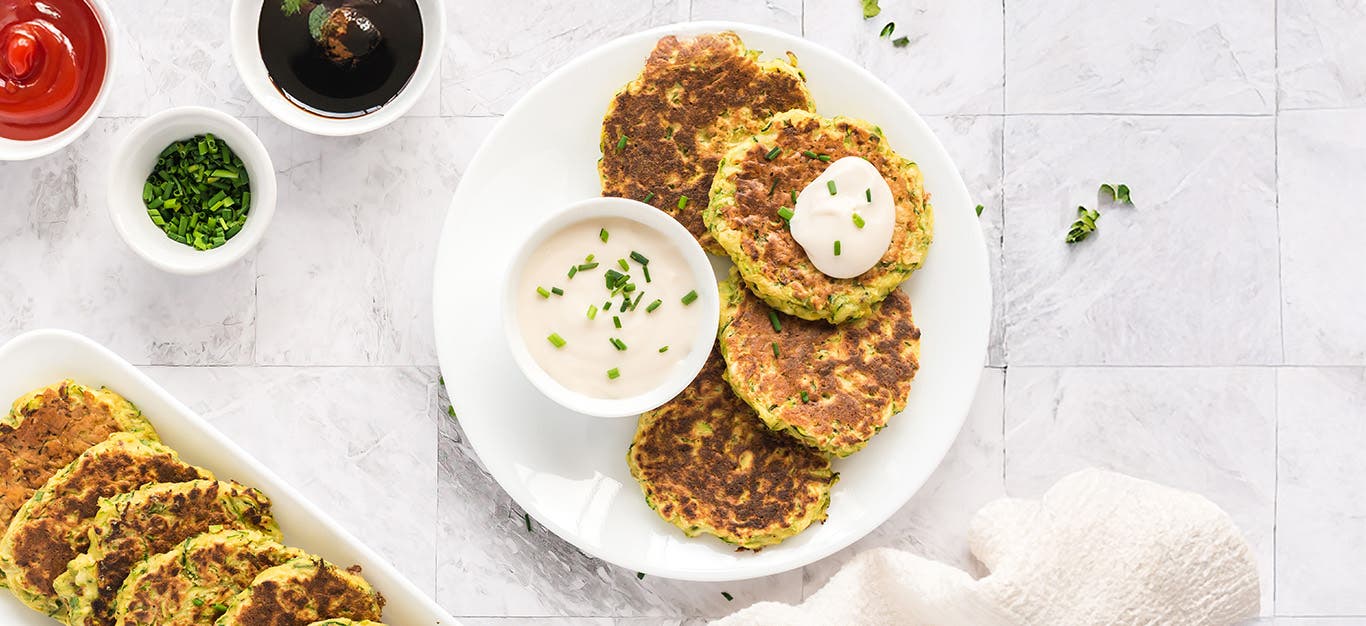 Zucchini Fritters on a white plate with a small white bowl of sauce, next to a larger serving plate of fritters
