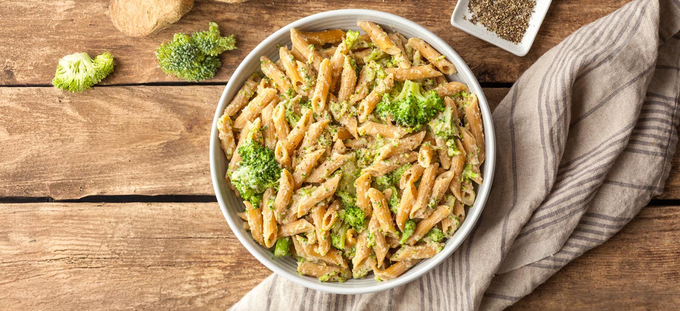 Big bowl of vegan celeriac mac and cheese with penne pasta and broccoli florets, on a rustic wooden table with a kitchen towel and seasonings to the side