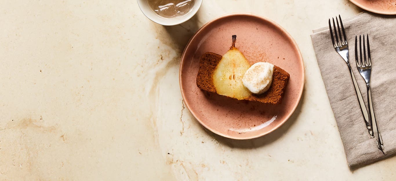 A slice of Gingerbread Loaf with Poached Pears on a pale pink plate, topped with a dollop of Aquafaba Whipped Cream.