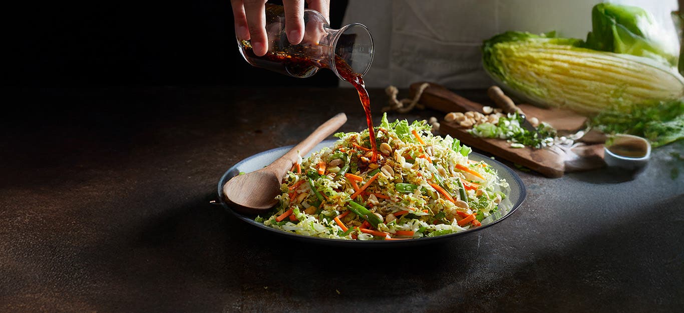 Napa Cabbage Salad in a bowl on a black counter, with a person pouring dressing over the top