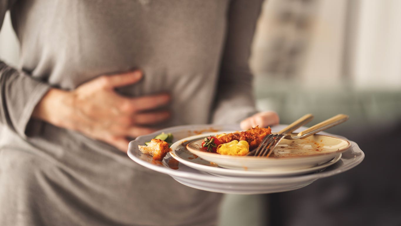 Person having heartburn and clutching their lower chest area with one hand while holding a plate of mostly eaten food in the other