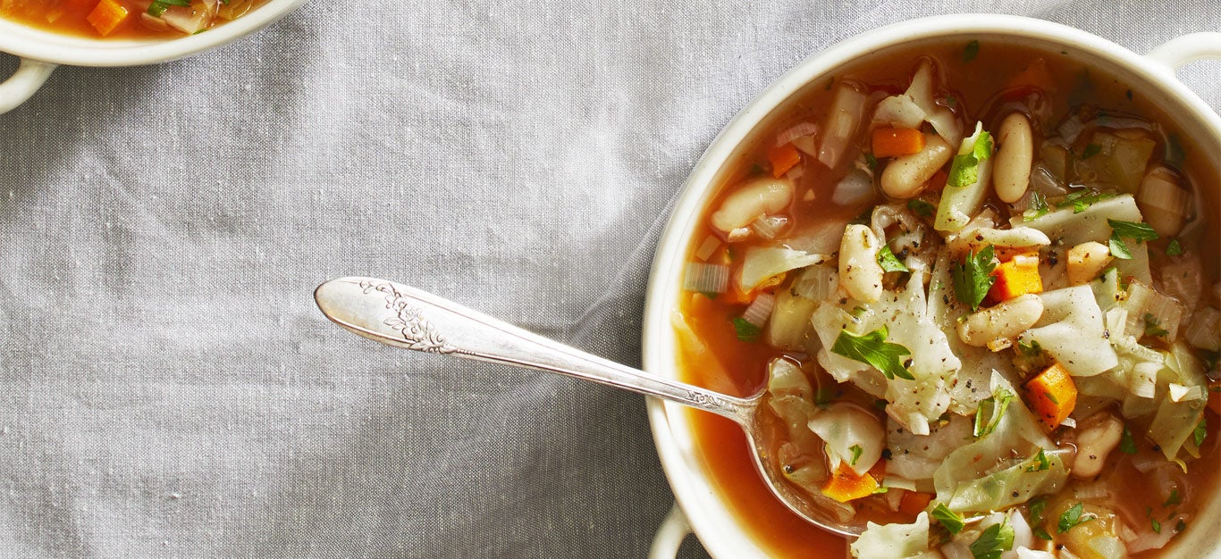 Green Cabbage and White Bean Soup in white ceramic bowls with a metal spoon against a gray cloth background