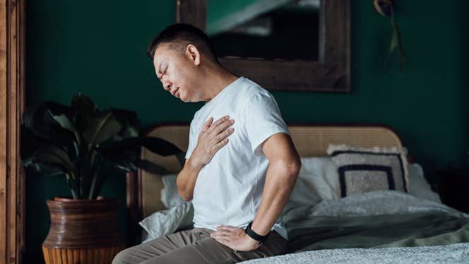 Middle-aged Asian man sitting on the edge of a bed, holding his chest in heartburn pain