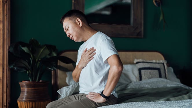 asian-man-suffering-from-heartburn Middle-aged Asian man sitting on the edge of a bed, holding his chest in heartburn pain