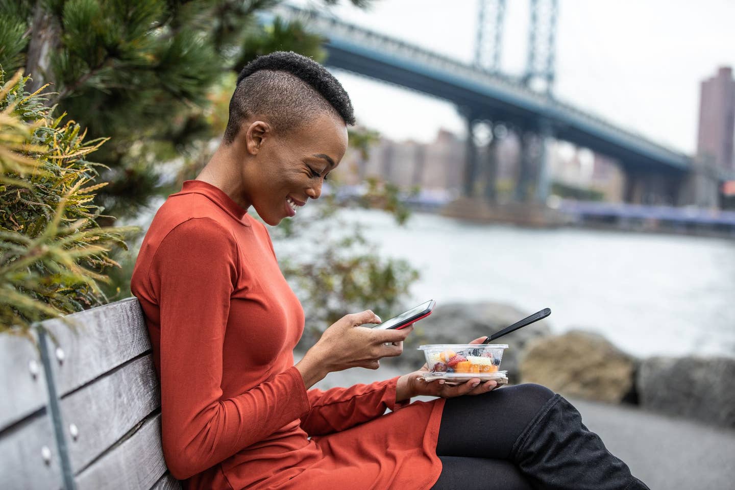 A young Black woman sits by the river in New York City eating healthy plant-based food