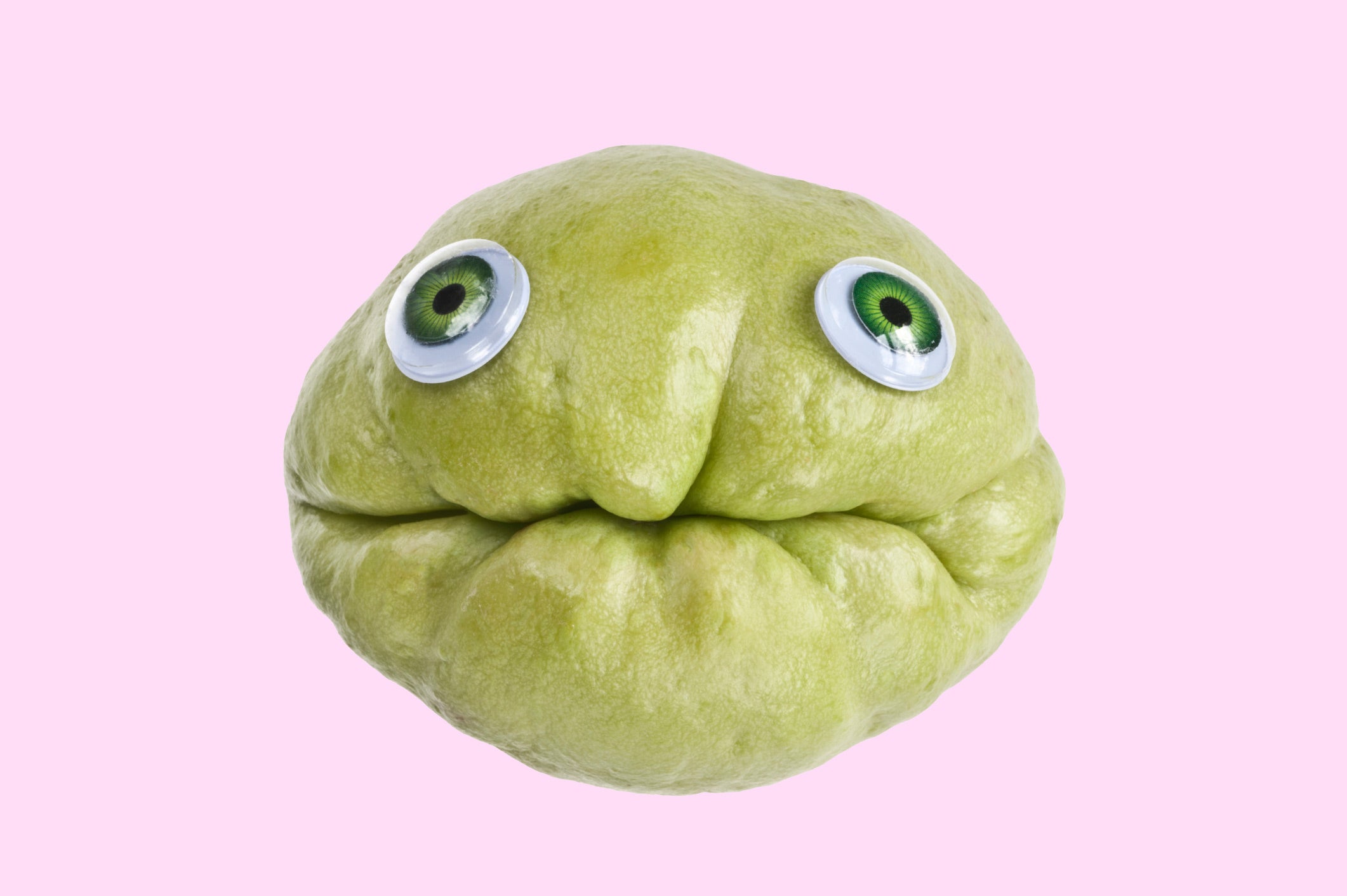 Funny pic of a chayote squash with googly eyes glued to it, angled so that the groove of the chayote looks like a mouth, on a solid pink background