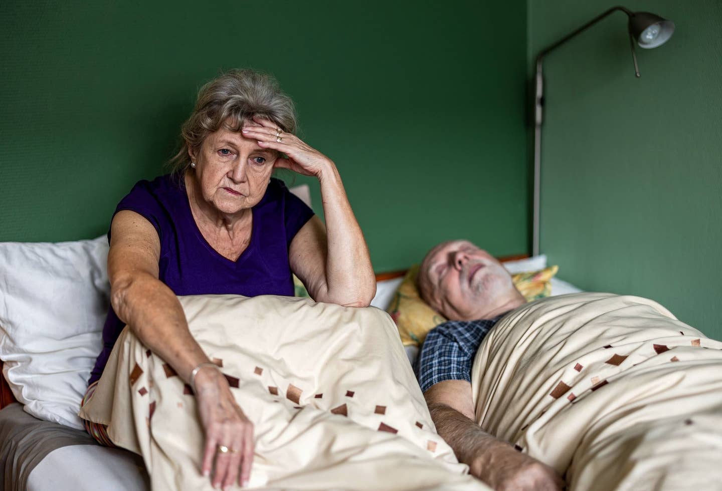Senior woman can’t sleep because her husband is snoring An older white woman sits up in bed, unable to sleep, while her husband lies snoring next to her