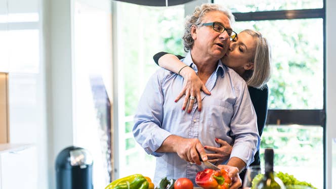 A senior man stands at his kitchen island chopping bell peppers, while a woman wraps her arms around him from behind and gives him a kiss on the cheek