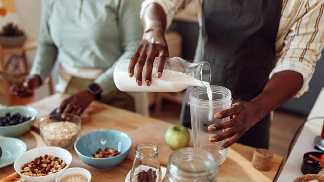 In a kitchen with a wooden countertop, a man and woman arrange bowls of healthy plant based ingredients, with the man pouring vegan milk into a smoothie blender