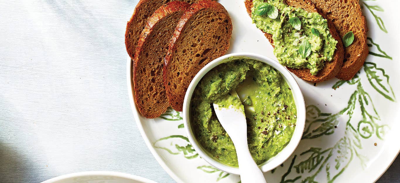 Pesto Artichoke Spread in a small white bowl with a serving knife on a plate with slices of whole wheat bread