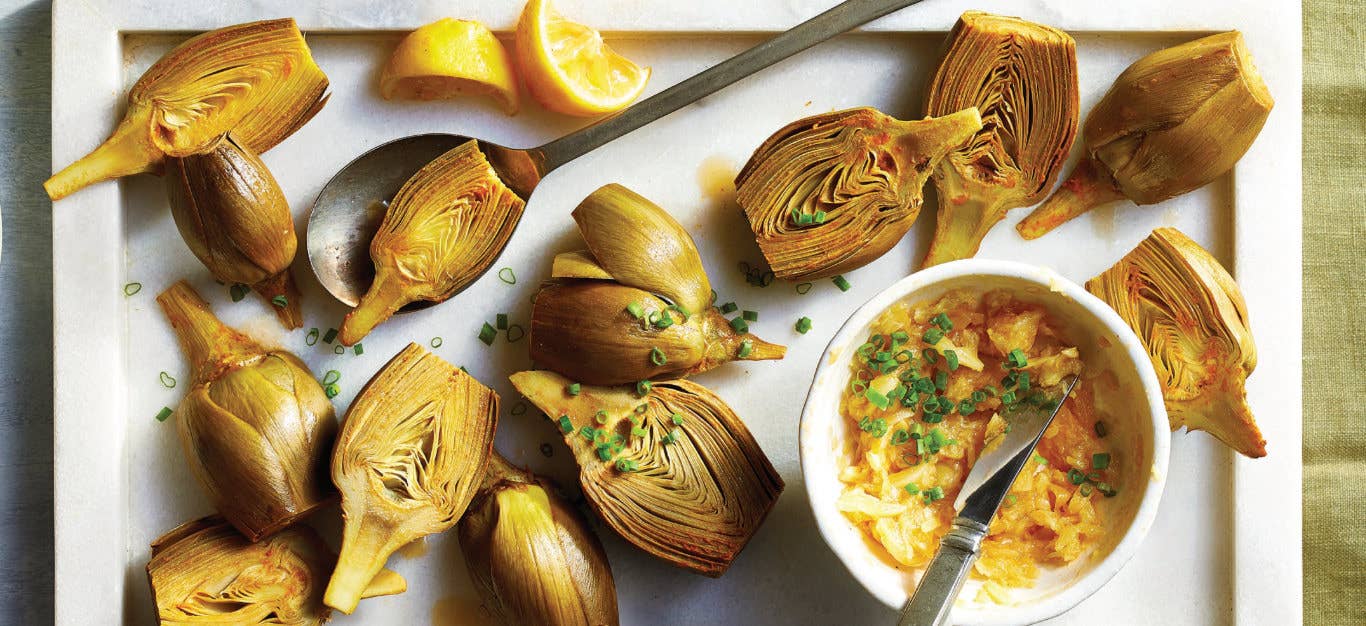 Lemon-Garlic Roasted Artichoke halves on a white platter next to a white ramekin of pressed garlic