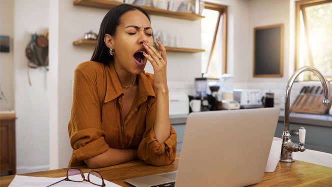 A dark haired woman sits at a kitchen counter with her laptop open and yawns from exhaustion