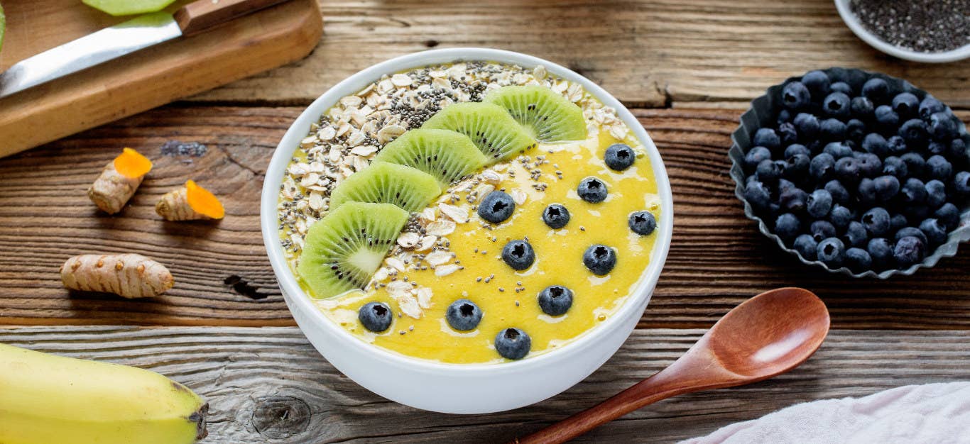 Mango Smoothie Bowl on a rustic wooden table next to a bowl of fresh blueberries, fresh turmeric root a banana and a wooden spoon