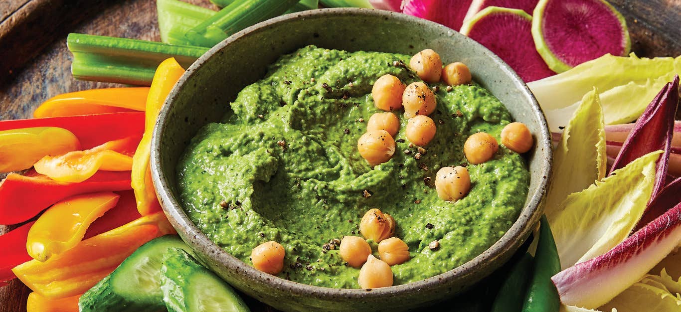 Fresh Spinach Hummus garnished with whole chickpeas on a round platter surrounded by bite sized cucumber, celery, radish, and bell pepper