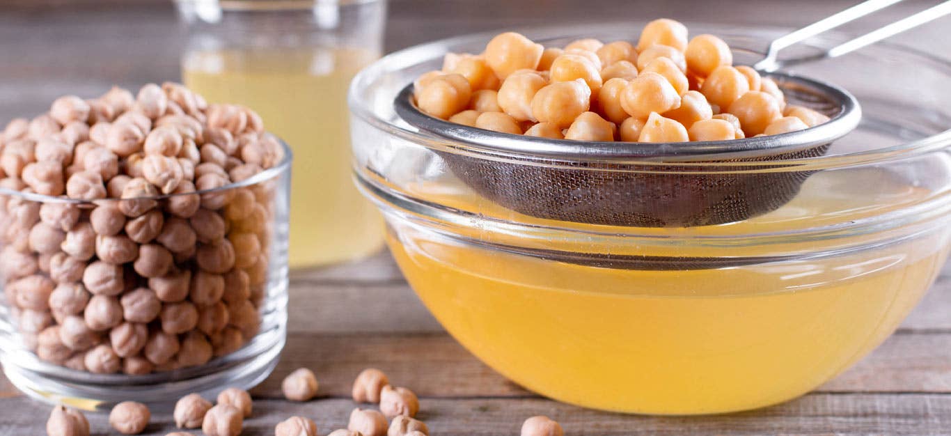 A mesh strainer full of chickpeas is placed over a glass bowl where aquafaba has drained down into it. A glass of dried chickpeas sits to the lefthand side.