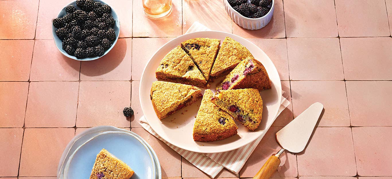 Blackberry Polenta Cake on a pink plate with one slice removed and on a nearby plate. There are two small bowls of fresh blackberries in the background