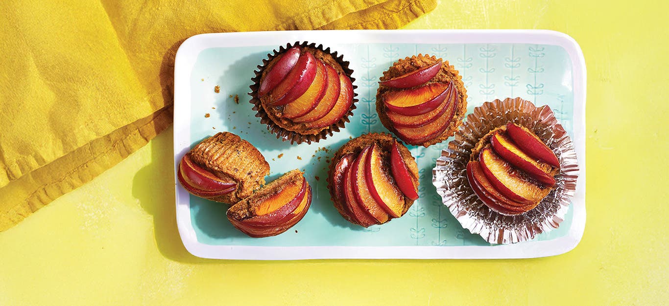 Fresh Plum Muffins in foil muffin papers on a pale blue and white plate with plum slices fanned across the top of each muffin