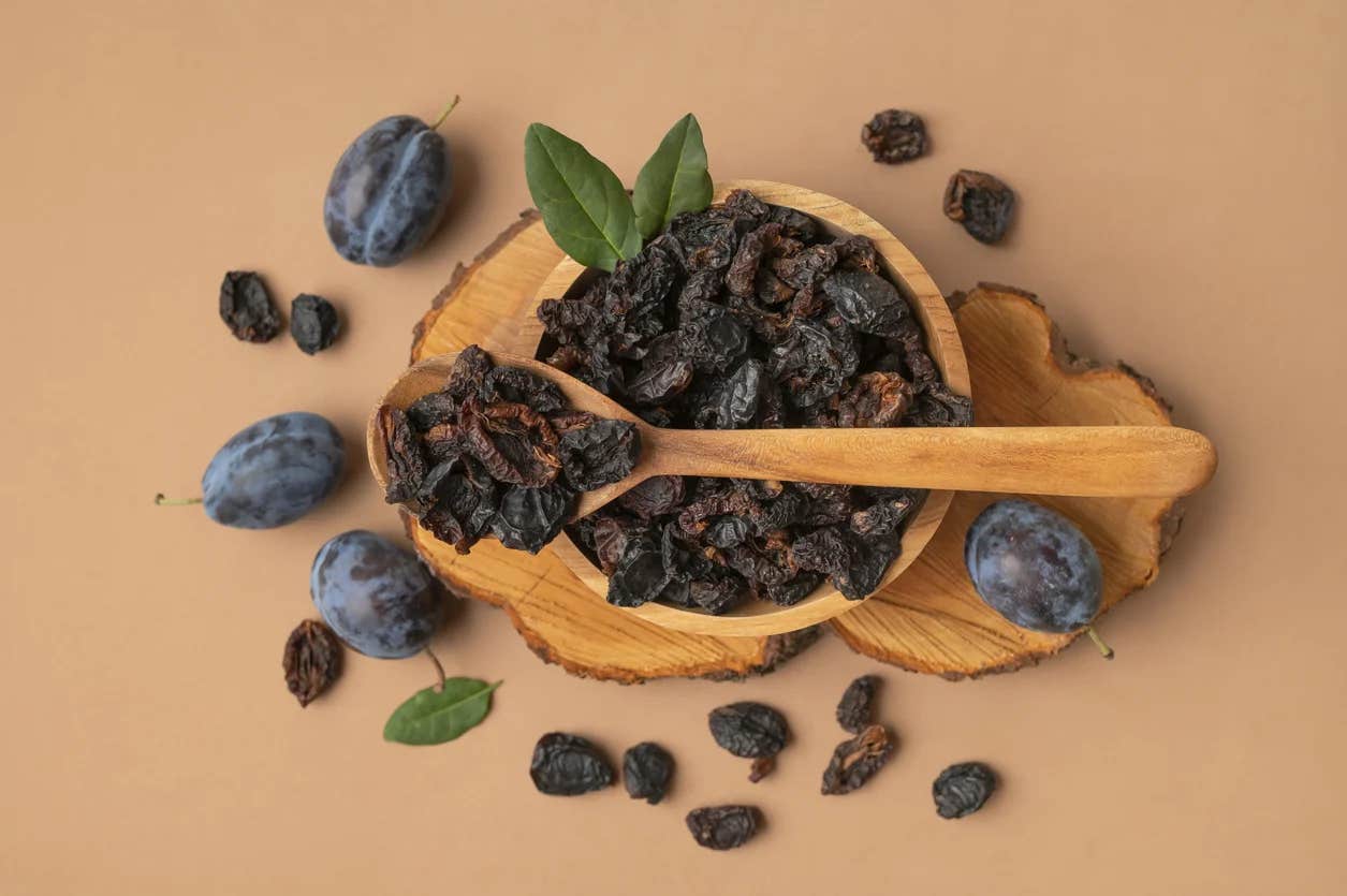 a bowl of prunes shown next to a few fresh plums on a wooden surface