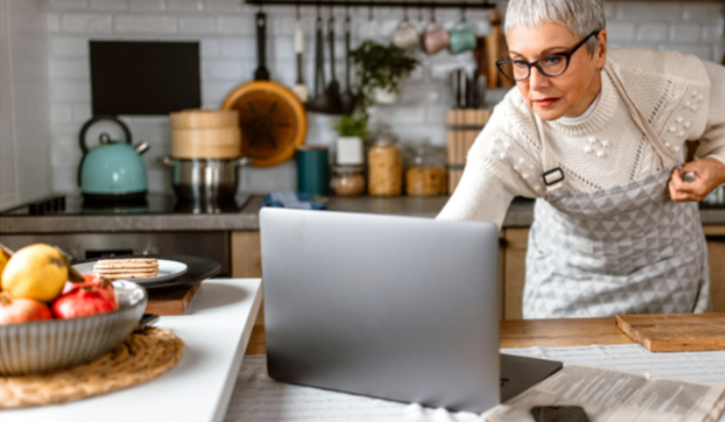 a senior woman wearing an apron stands leaning over a laptop in a well-equipped kitchen