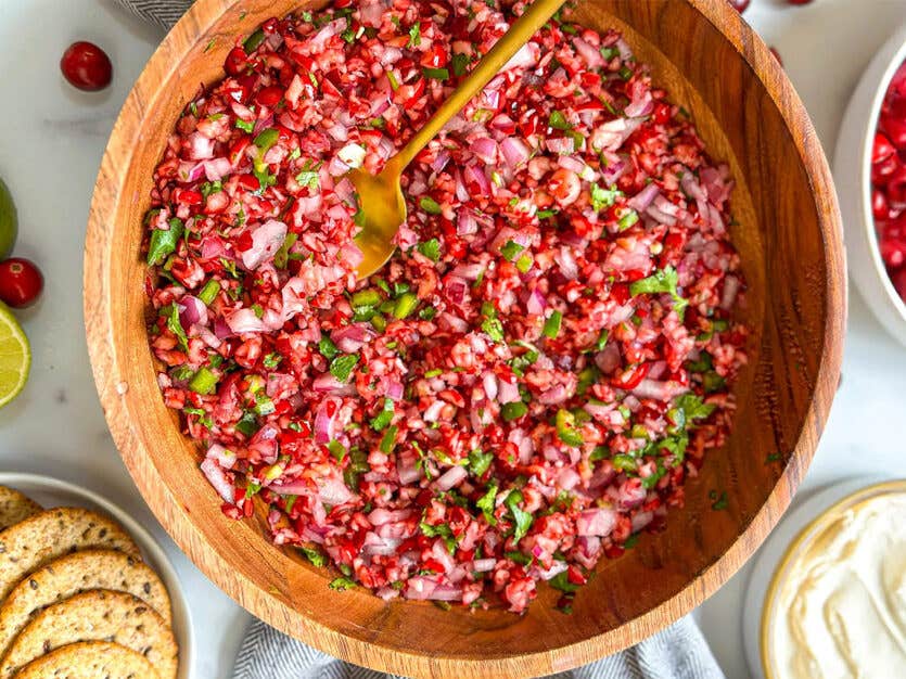 Jackie Akerberg’s Cranberry Salsa in a wooden serving bowl with a copper spoon, beside limes and crackers and vegan ricotta
