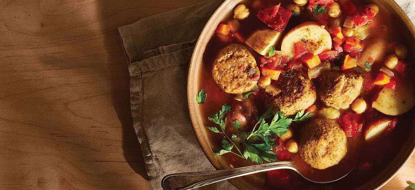 A rustic brown serving bowl of Vegan Kefta Tagine (Moroccan Meatballs) on a brown cloth napkin, with a spoon in the bowl