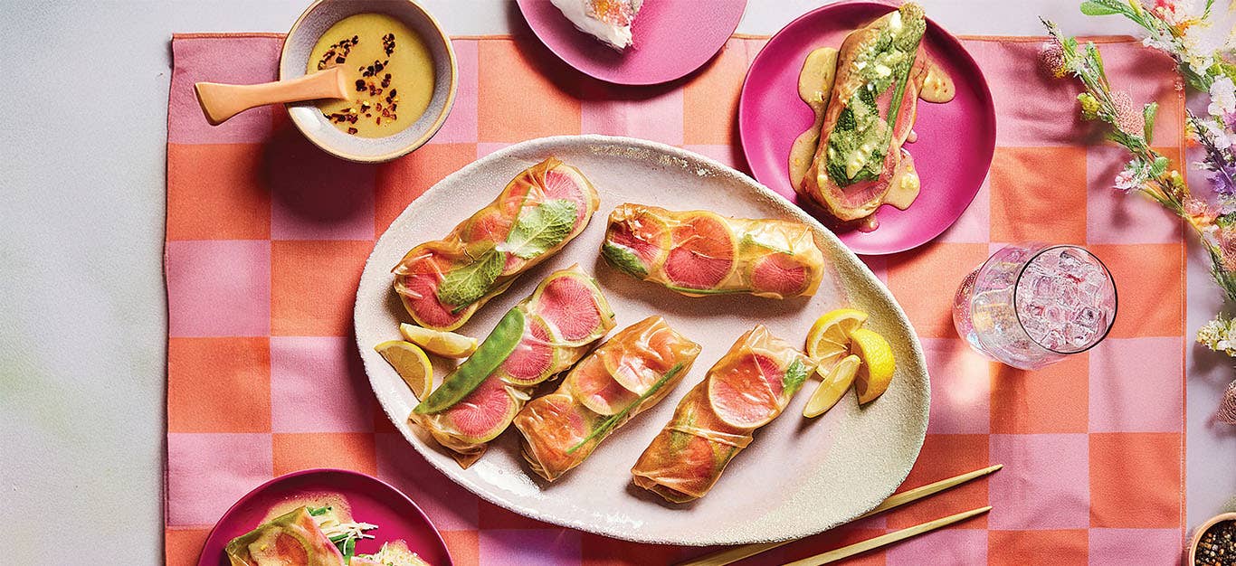 Colorful Watermelon Radish Spring Rolls on a white platter on a pink and orange checked placemat. There is a small bowl of Lemon-Ginger Dipping Sauce and two bright pink serving plates with one roll on each, smothered with sauce.