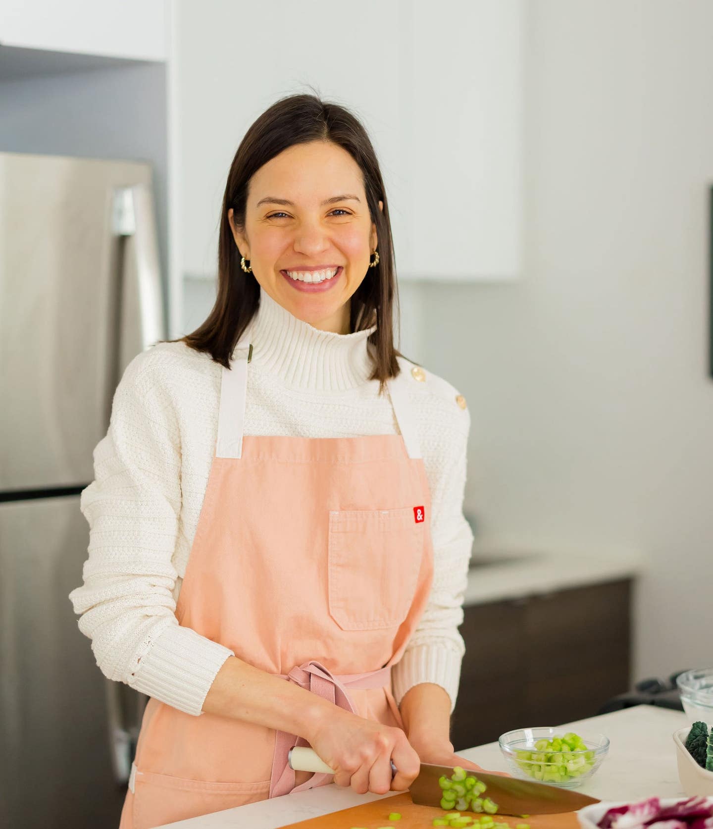 Registered dietitian and cookbook author Gena Hamshaw stands in her kitchen wearing an apron and smiling as she slices scallions