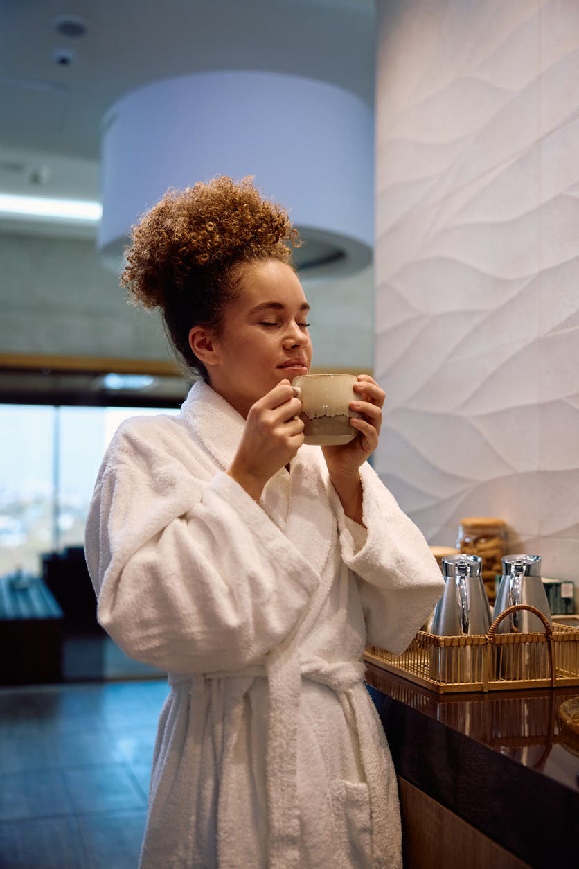 Woman wearing a robe at a spa enjoys a cup of herbal tea with her eyes closed