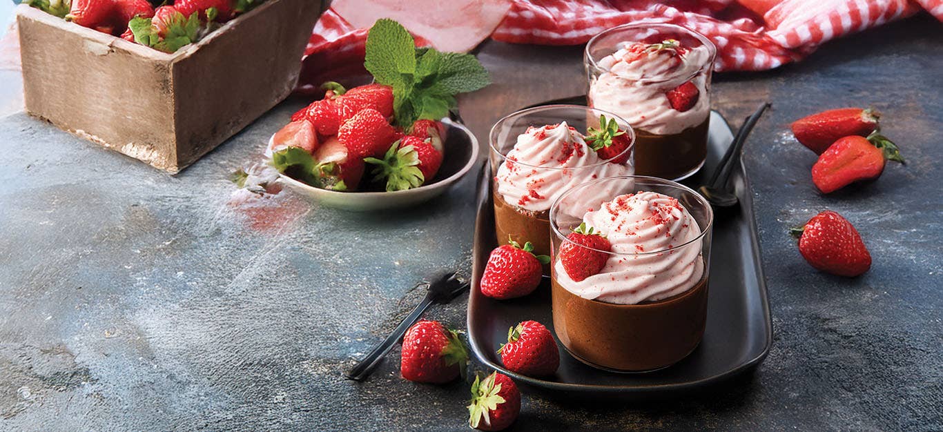 Three glass parfaits of Black Bean Chocolate Mousse with Whipped Strawberry Cream on a small tray, with fresh strawberries scattered