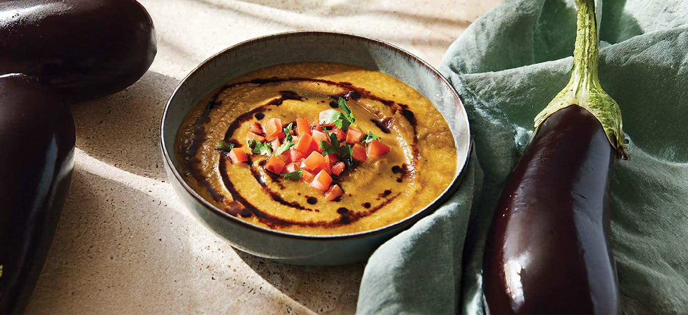 A black bowl of orange-hued soup topped with a drizzle of balsamic glaze next to a whole globe artichoke