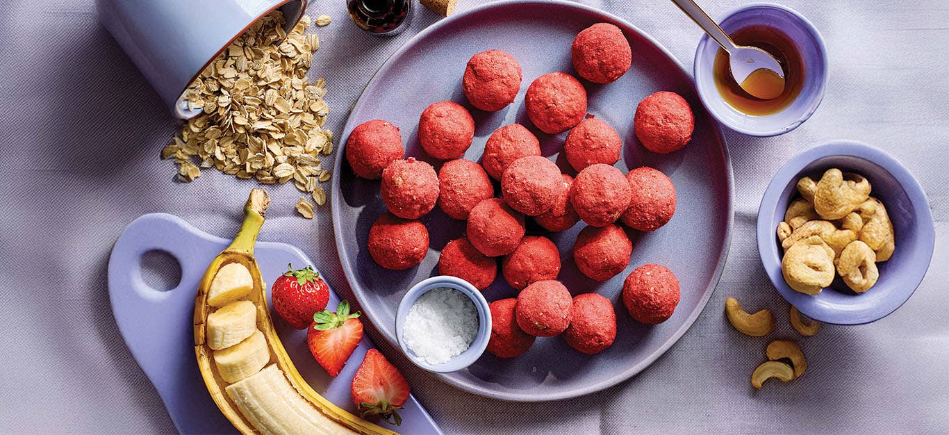 Strawberry Bliss Bites on a blue plate with small containers of cashews, rolled oats, and a banana, nearby