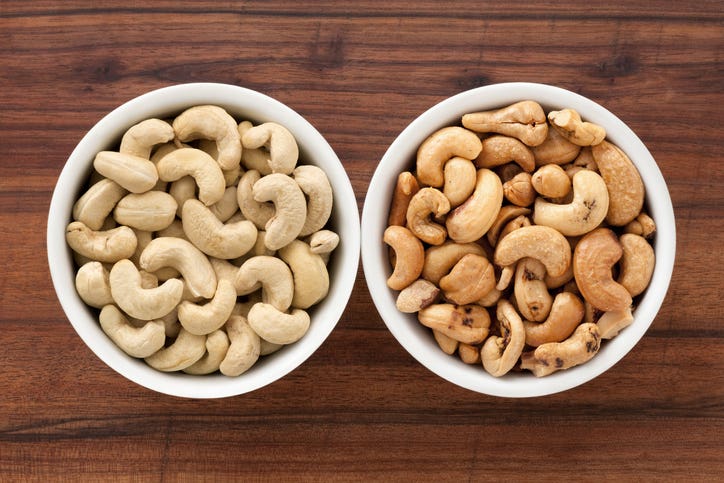 Top view of side by side bowls of raw cashews next to roasted cashews, which are darker