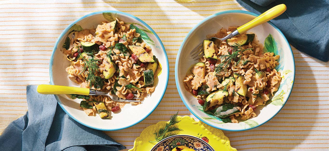 Two bowls of Charred Lemon and Zucchini Pasta with a yellow-handled fork in each and two blue napkins on a yellow and white striped table cloth