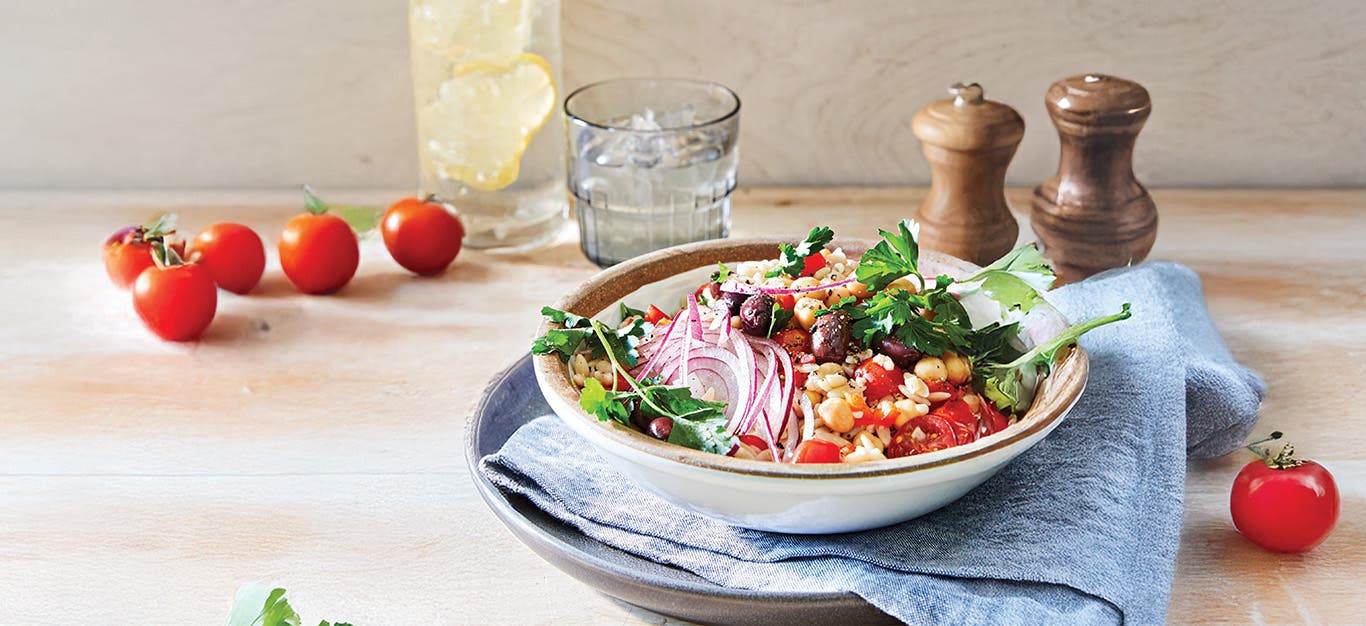 A bowl of Lemon Orzo Salad garnished with red onion and fresh parsley, with cherry tomatoes dotted on the table around the bowl