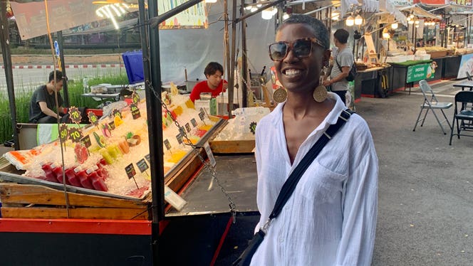 Vegan travel expert Tracye McQuirter stands in front of a fruit stand at an outdoor market overseas