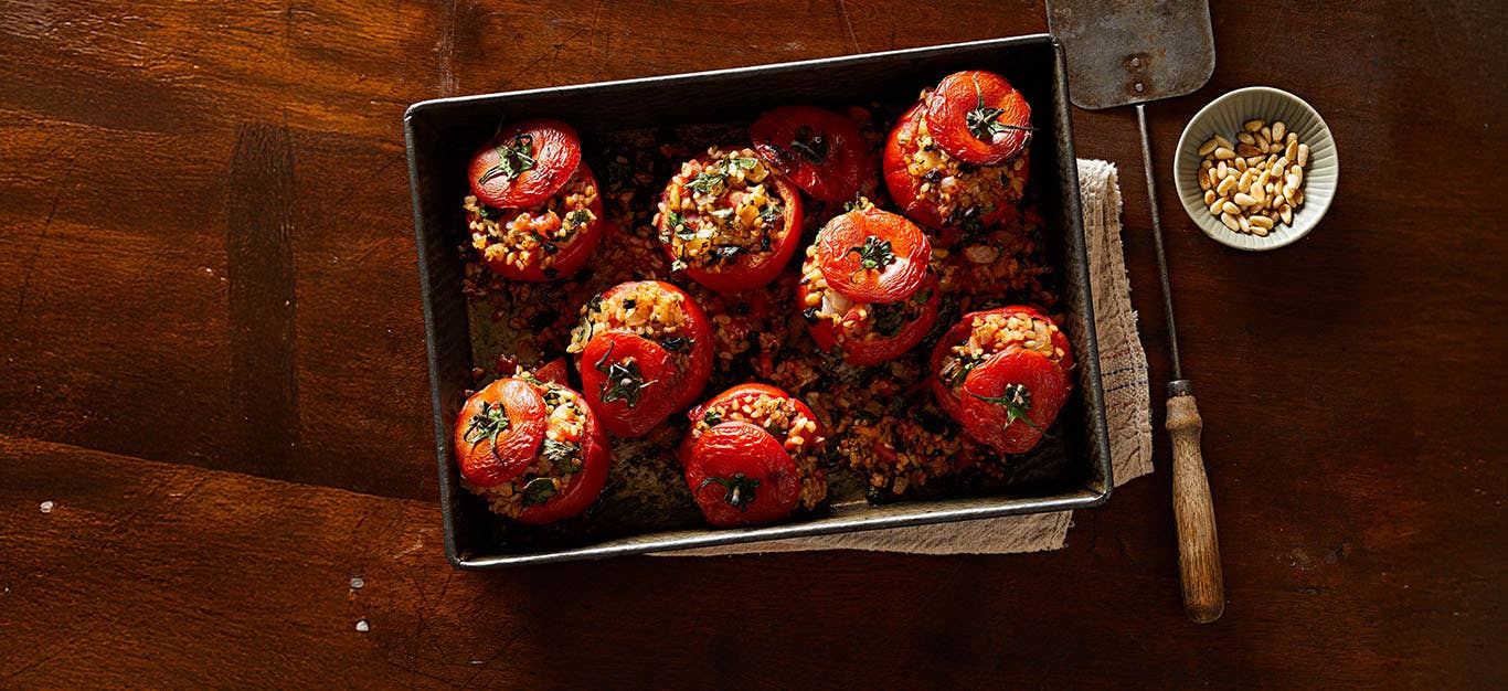 Greek-Style Stuffed Tomatoes on a baking tray with a small bowl of pine nuts to the side