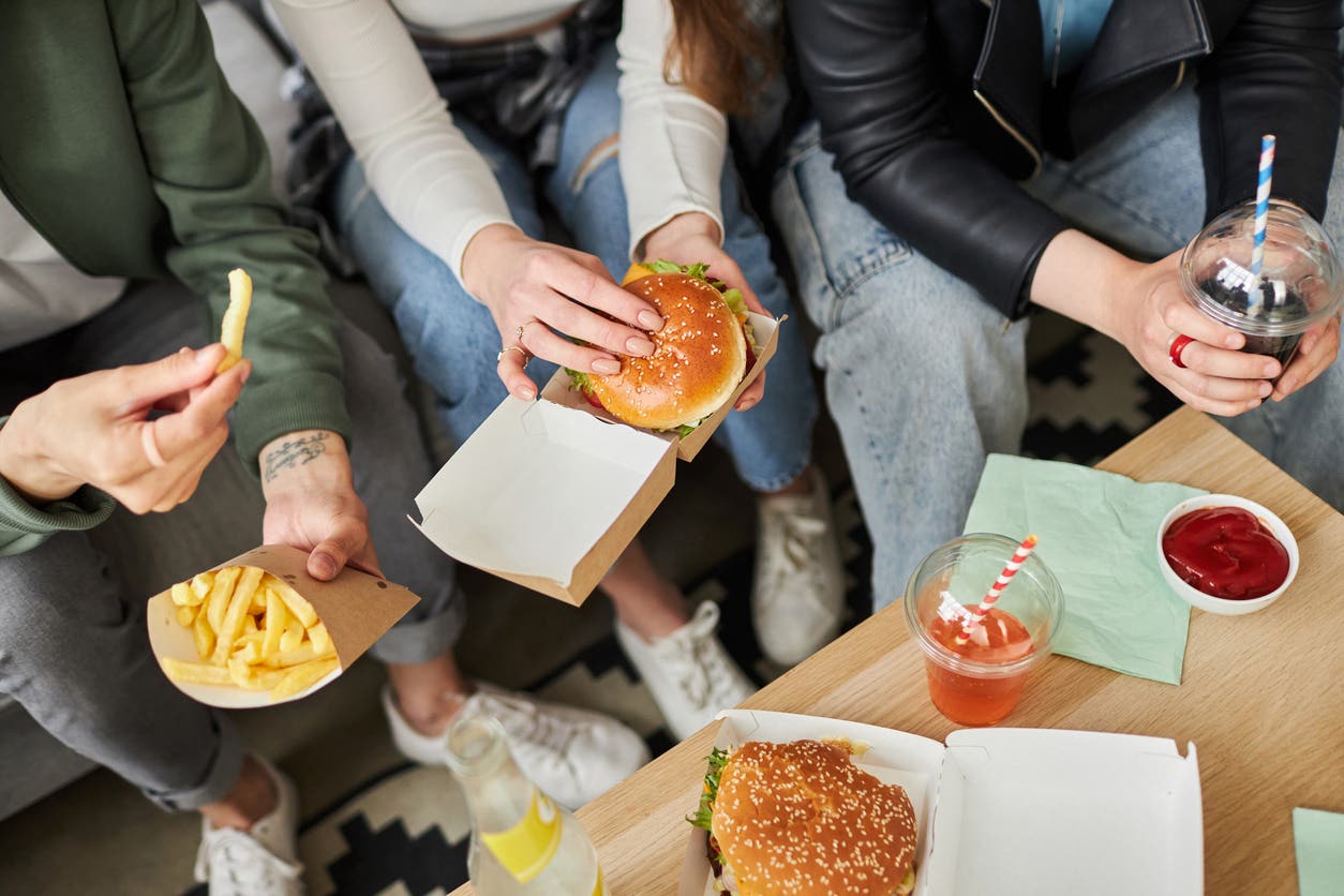 Young people sit around a coffee table eating ultraprocessed fast-food including cheeseburgers, French fries, and frozen soft drinks