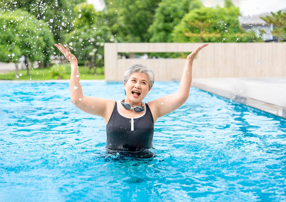senior-woman-splashing-pool A senior woman stands in a pool outside looking joyous with her arms raised upward, wearing an athletic swimsuit and goggles around her neck