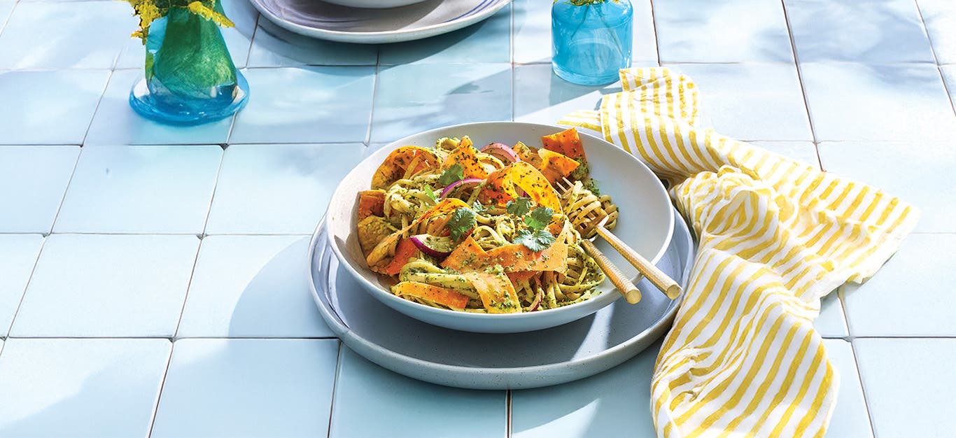 A bowl of Fettuccine with Spinach Pesto and Carrot Ribbons in a pale blue bowl on a blue plate on pale blue tiles with a yellow and white striped unfolded napkin next to the plate