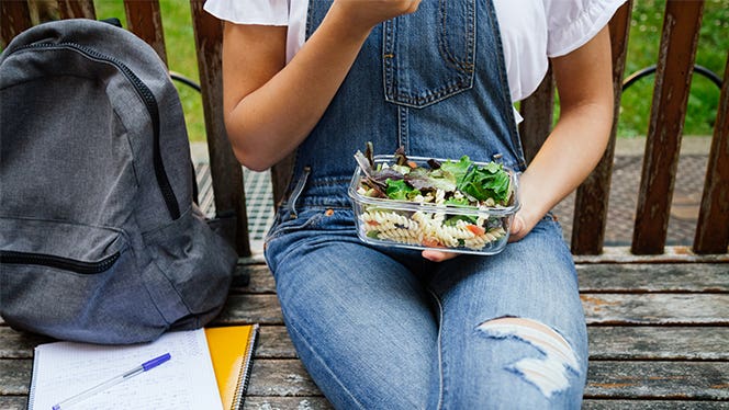Our Favorite Lunchboxes for Kids and Adults Unidentified woman in overalls sitting on a park bench eating her lunch, with her backpack next to her