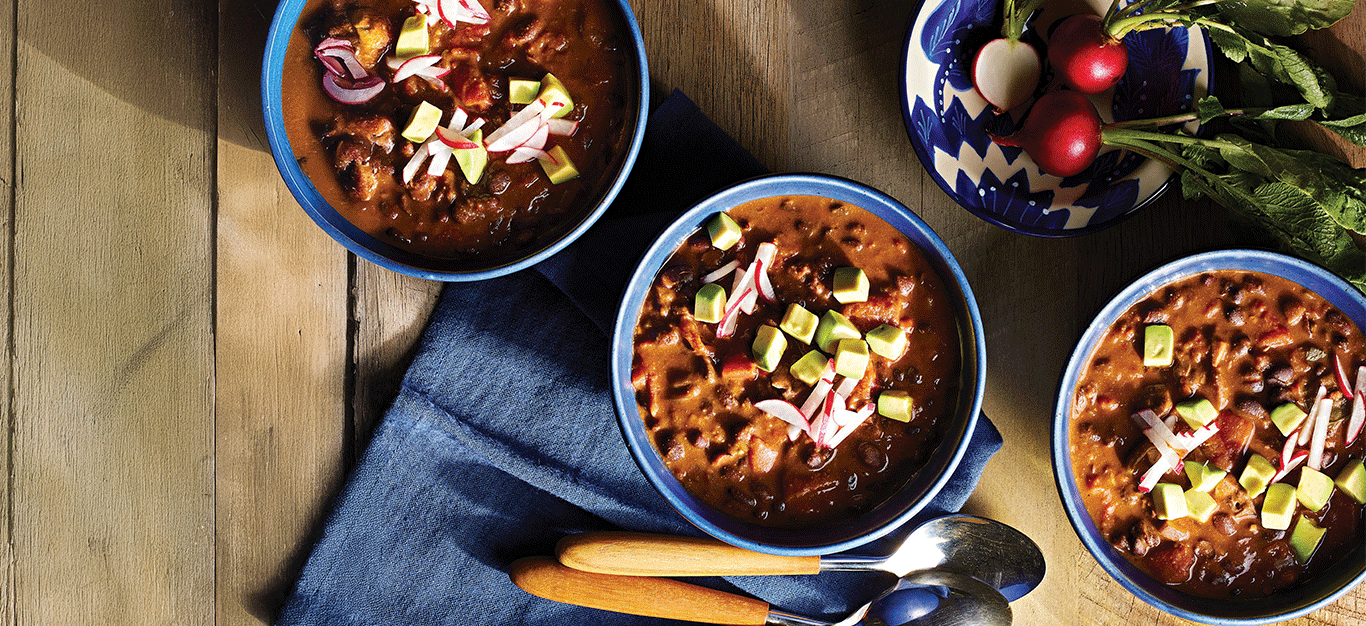 Three bowls of Mole Chili on a wooden table, with a blue napkin, wooden-handled spoons, and blue cermamic bowls