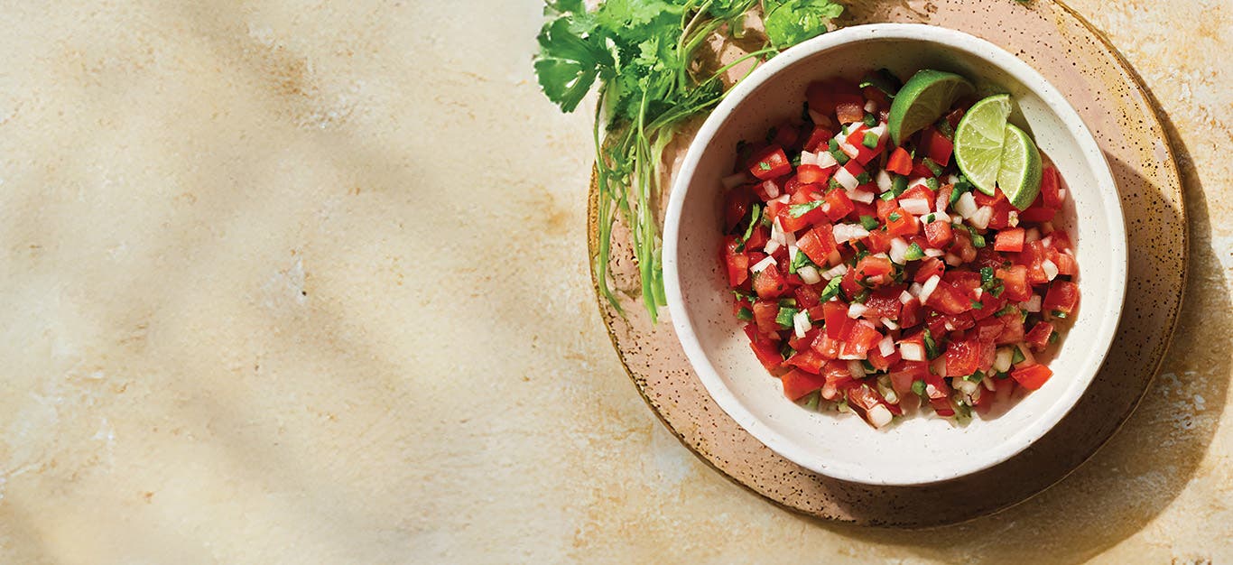 Pico de Gallo in a pale pink bowl on a brown ceramic plate, with cilantro on the side, and lime wedges in the bowl