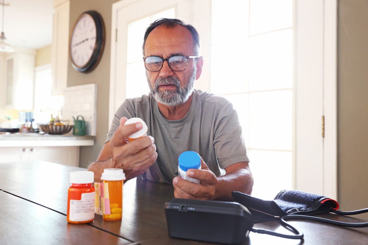 A senior citizen man sits at his dining room table sorting through bottles of prescription medications as sunlight filters in through the window behind him bathing the room with a soft glow of light. A blood pressure machine rests on the table in front of him.