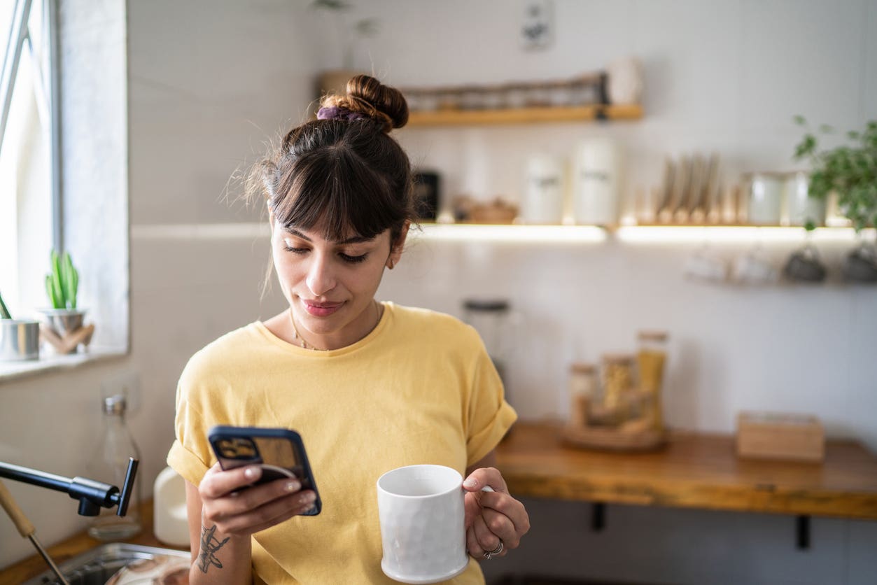 Young woman wearing a yellow shirt looking at phone and texting in kitchen while holding a mug