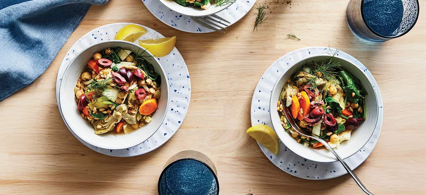 Two white serving bowls on white plates with lemon wedges on the side. Each bowl is full of a colorful mix of Greek-Style Farro with Chickpeas and Artichokes