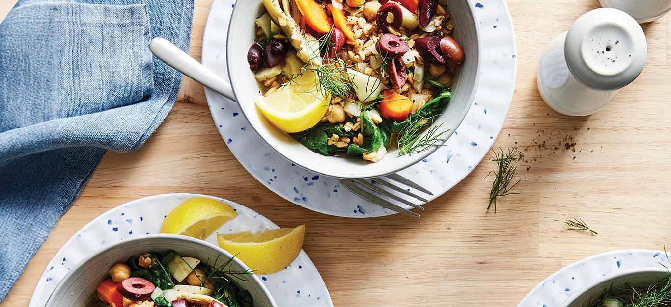 Two white serving bowls on white plates with lemon wedges on the side. Each bowl is full of a colorful mix of Greek-Style Farro with Chickpeas and Artichokes
