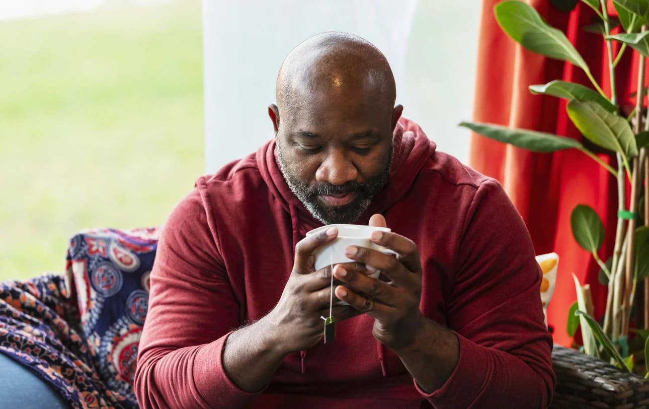 iStock-2173201500 copy A middle-aged man sits in a cozy brightly decorated living room near a window with a mug of tea in his hands
